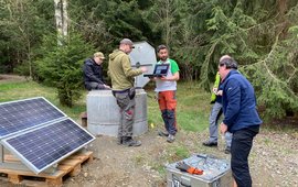 Fünf Menschen stehen um ein Bohrloch, das mit Beton abgeschlossen ist. Ein Solarmodul liefert Strom für Messgeräte. Im Hintergrund: Wald.