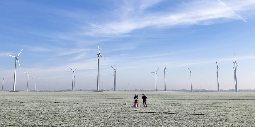 Einige Windräder im flachen Land. Davor stehen zwei Menschen auf dem bereiften Feld. Blauer Himmel mit Schleierwolken.